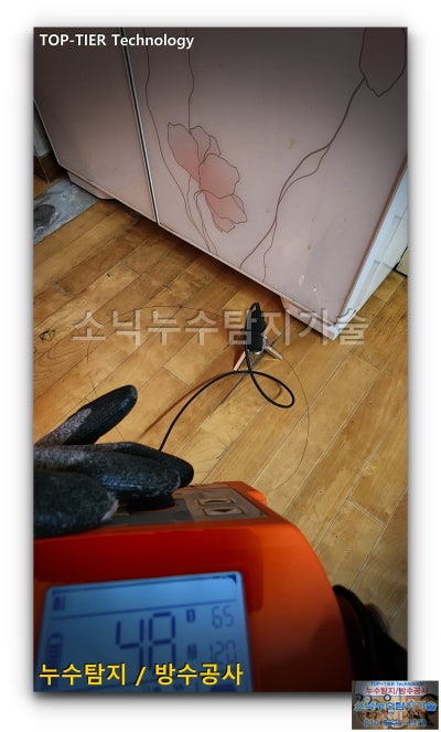 마루 바닥에 청음 센서를 올려놓고 주황색 본체 계기판 수치를 확인하는 수성구 아파트 누수 청음탐지 장면