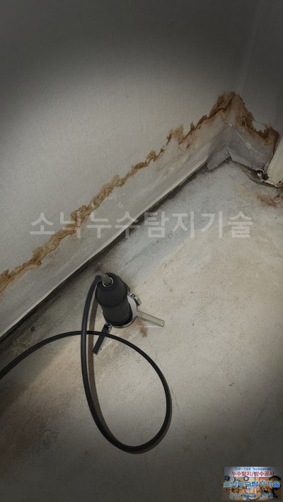 작은방 벽 아래 번진 물 자국 옆 바닥에 청음 탐지 장비를 올려놓고 누수음을 듣는 모습